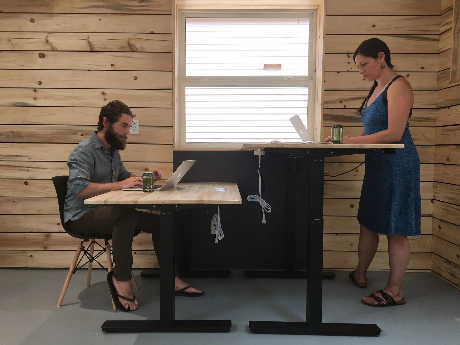 Two members at adjustable sit-stand desks in The Coop, one seated and one standing, against a wood-paneled wall.