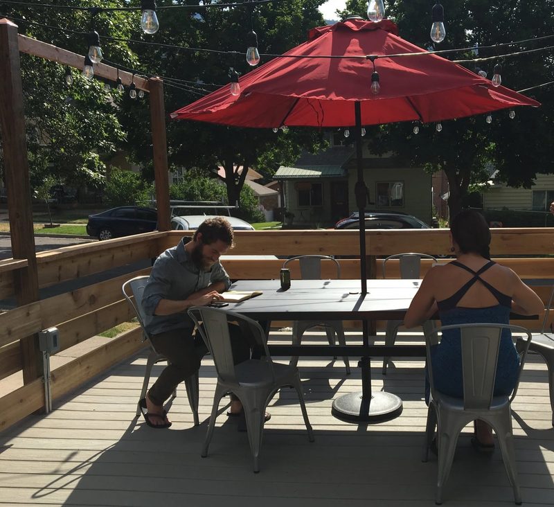 Two members working at a table on The Coop's outdoor deck under a red umbrella, with string lights overhead.